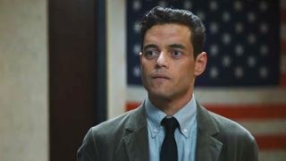 rami malek wears a tie and stands in front of the american flag in a still from the amateur