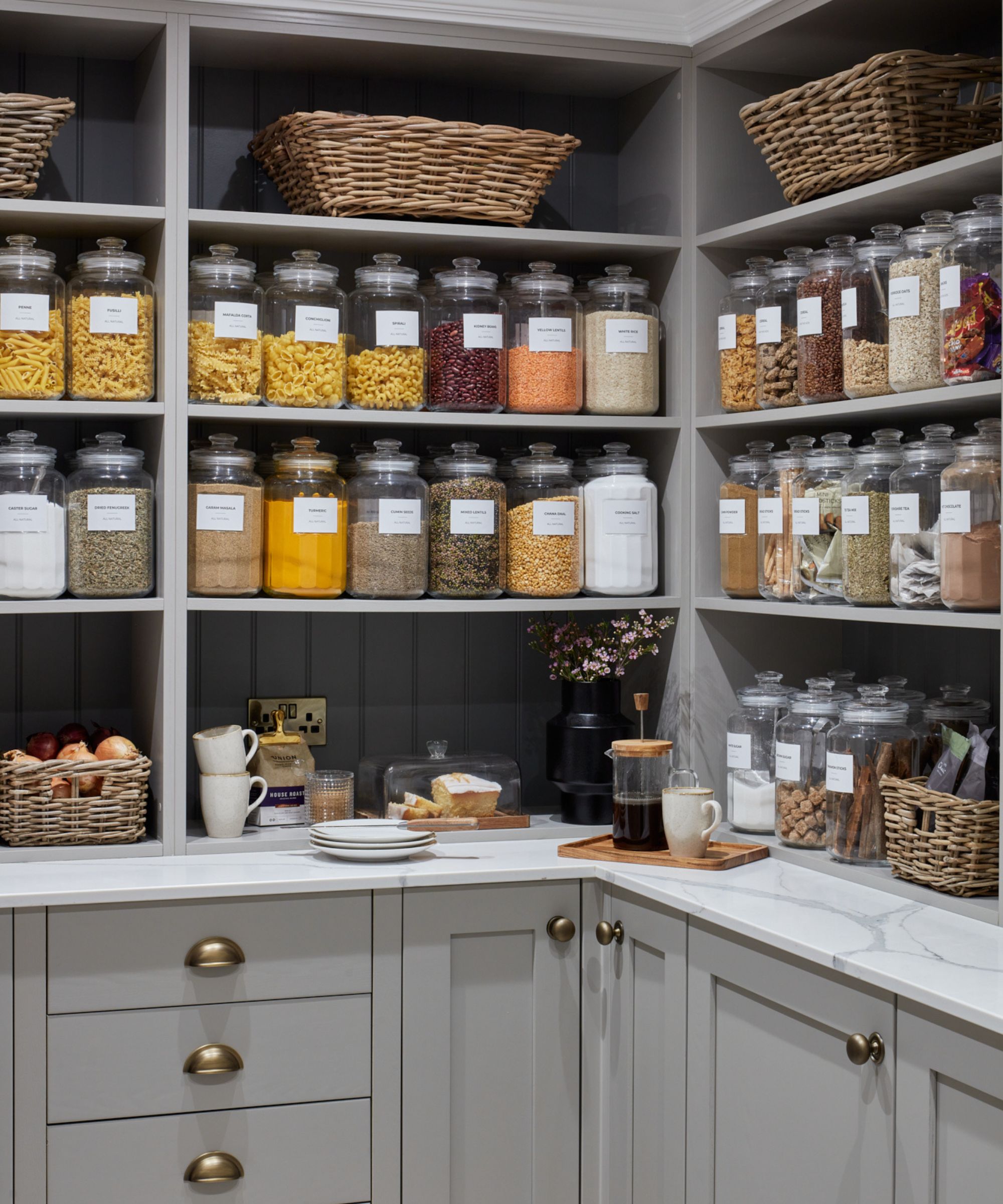 A neatly organized pantry with grey cabinets, brass knobs, marble countertops, glass jars labeled with dry goods, wicker baskets, a coffee setup, and a small display of flowers