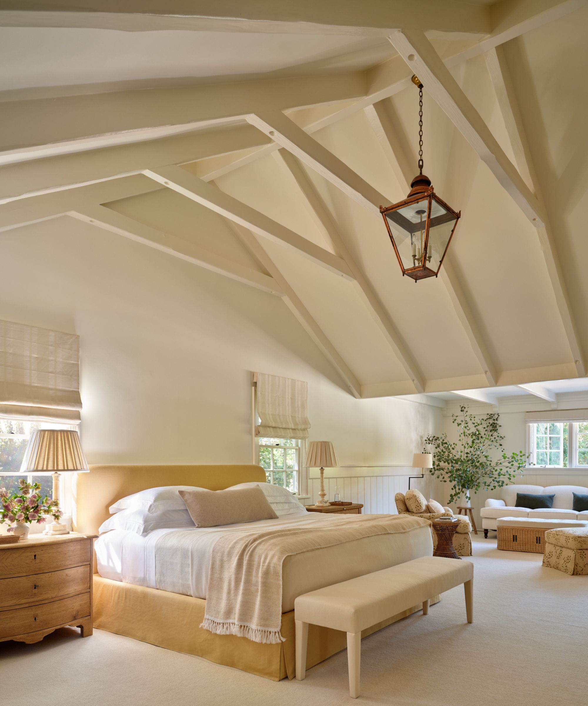 a large neutral primary suite with vaulted ceiling and a yellow upholstered bed, neutral linens, wooden nightstand and a cozy sitting area