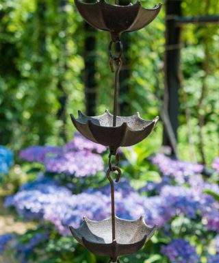 A silver umbrella rain chain with blue and purple hydrangeas with green trees behind it