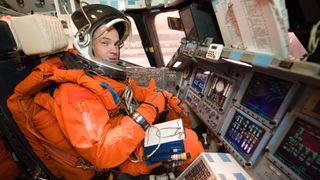 cj sturckow in an orange training suit sitting inside a space shuttle training cockpit with training materials on his knees