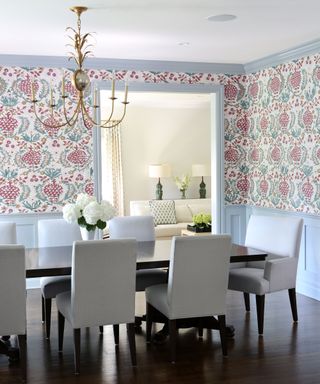 Light and bright dining room with floral wallpaper on the walls, wooden wall paneling painted a light blue shade, dark wood rectangular dining table with upholstered chairs, dark wooden flooring, metallic chandelier hanging over table