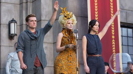 From left to right: Josh Hutcherson raising his hand in a Mockingjay symbol, Elizabeth Banks talking into a mic and Jennifer Lawrence raising her hands in Catching Fire. 