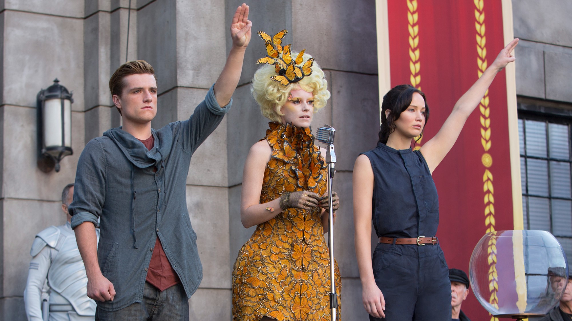 From left to right: Josh Hutcherson raising his hand in a Mockingjay symbol, Elizabeth Banks talking into a mic and Jennifer Lawrence raising her hands in Catching Fire. 