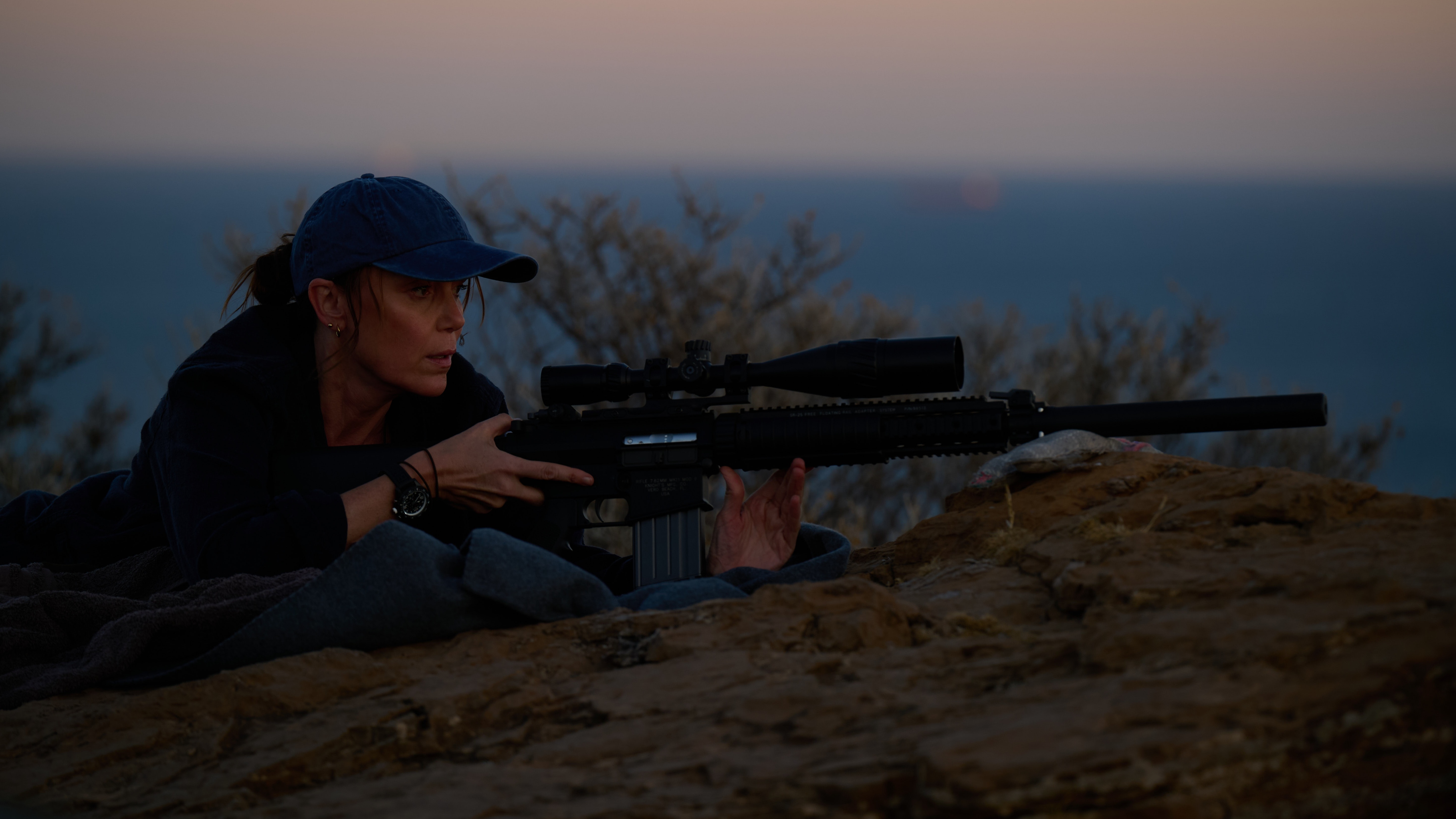 Julie lies on a sand bank with her gun ready to shoot as the sun sets behind her in The Assassin