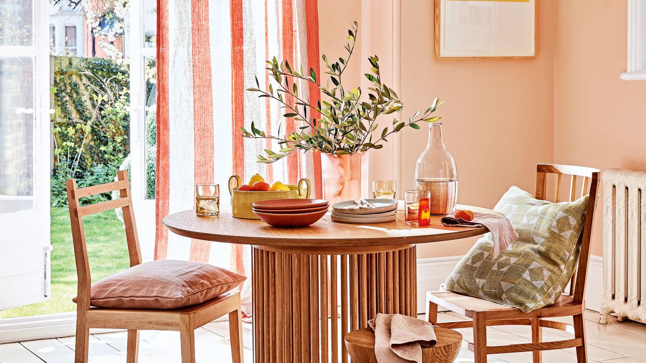 Round wooden dining table with two chairs, dressed with stoneware and olive sprigs in a glass vase and a white and orange striped curtain at an open door behind