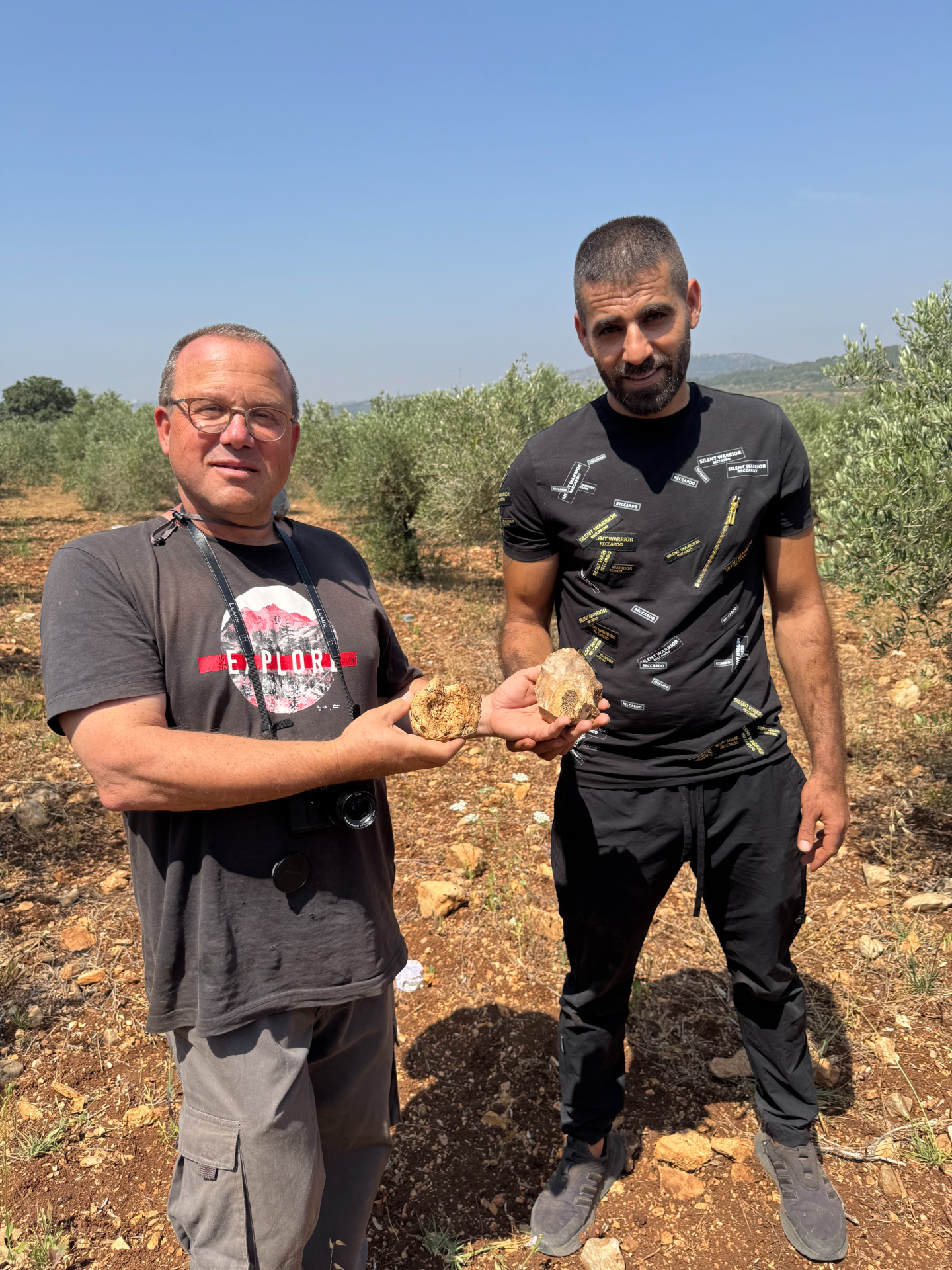 Two men wearing dark shirts and pants hold up two brown, round stones in a field.