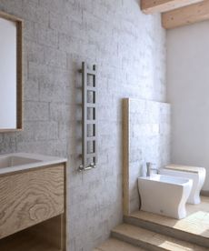 A grey ladder radiator on a stone bathroom wall, beside a wooden basin unit with a square mirror above, and some small steps up to a bidet and toilet. 