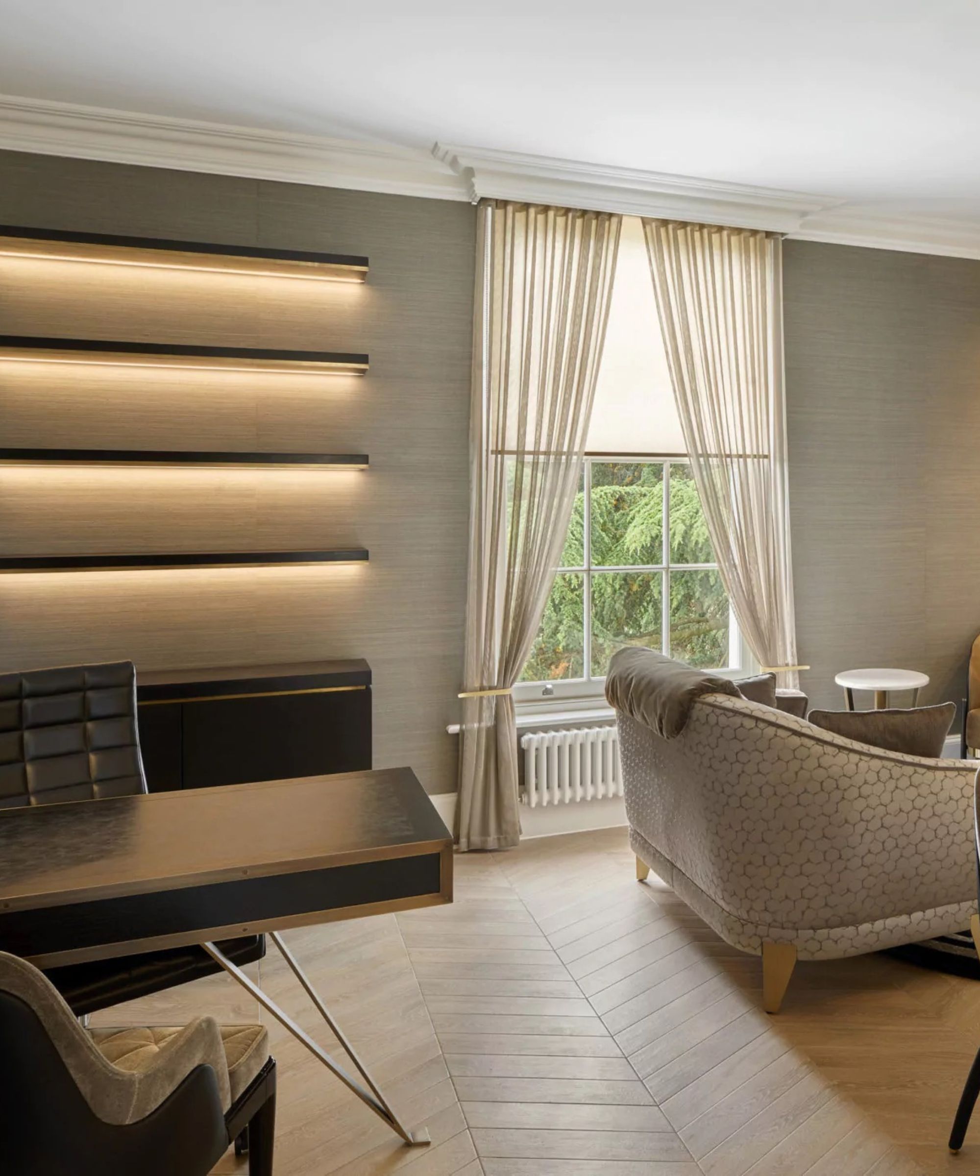 A stylish home office or lounge area with a sleek, modern design. On the wall, three horizontal floating shelves glow with soft integrated lighting. In front of the wall is a dark wood desk with slim metal legs, paired with a leather office chair.
