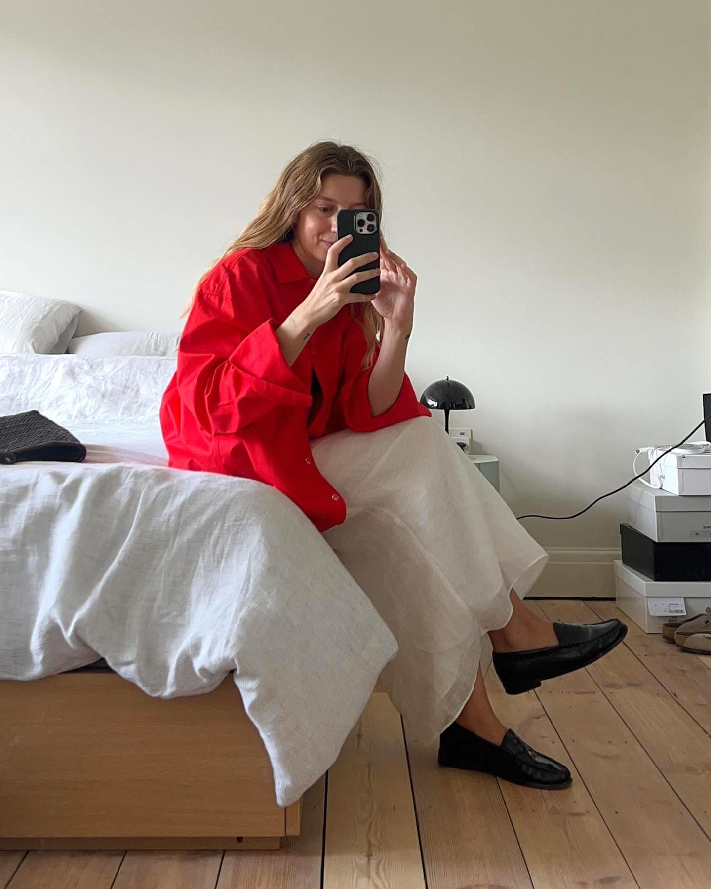 female fashion influencer Brittany Bathgate sits on her bed and poses for a mirror selfie with her iPhone wearing a bright red button-down shirt, gauzy white skirt, and black loafers