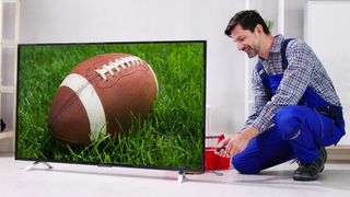 A man on coveralls is working on installing a TV. The TV is showing an American football on a grassy field