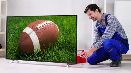 A man on coveralls is working on installing a TV. The TV is showing an American football on a grassy field