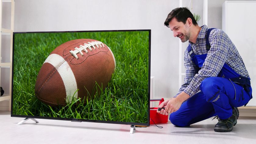 A man on coveralls is working on installing a TV. The TV is showing an American football on a grassy field