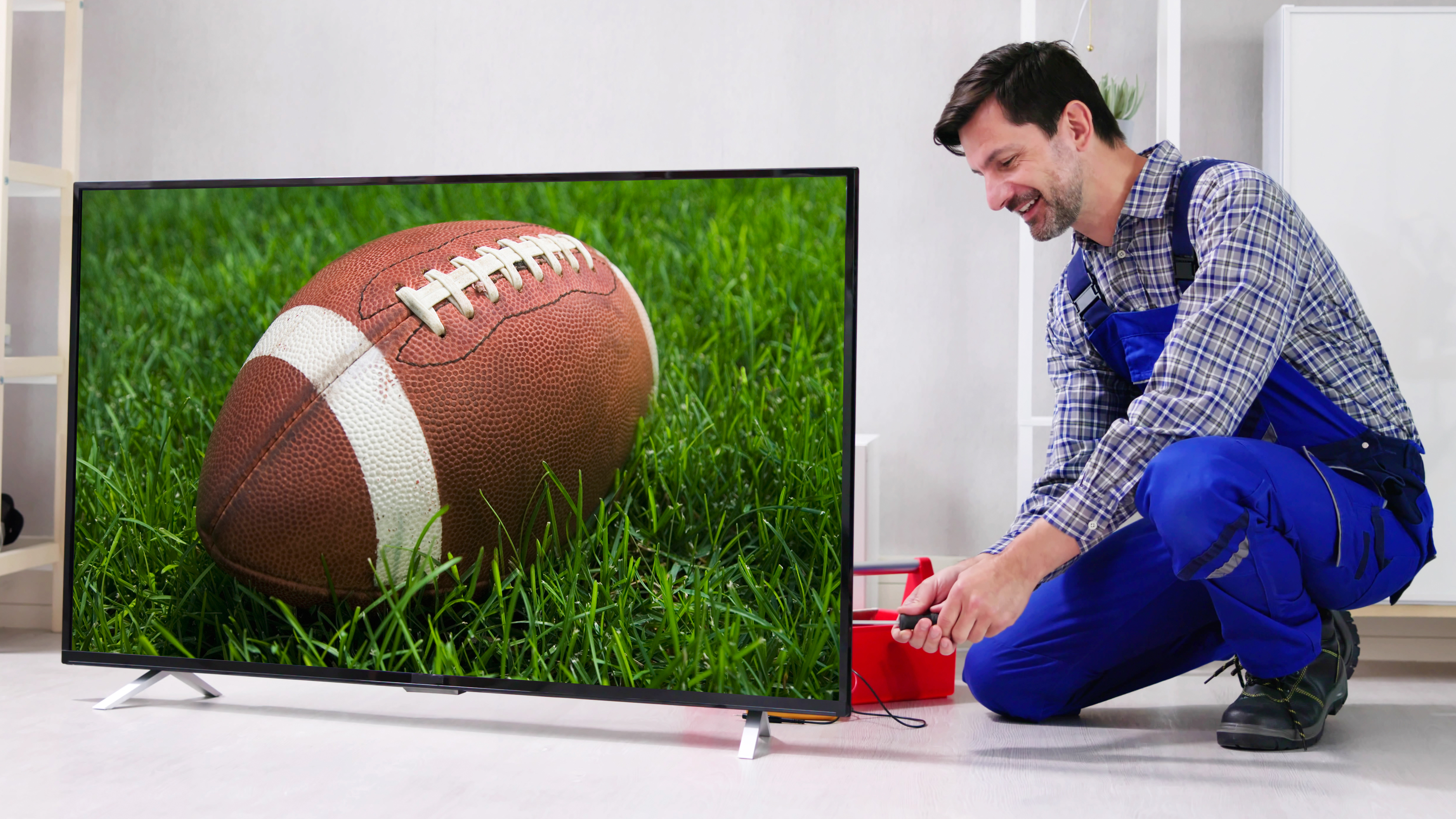 A man on coveralls is working on installing a TV. The TV is showing an American football on a grassy field