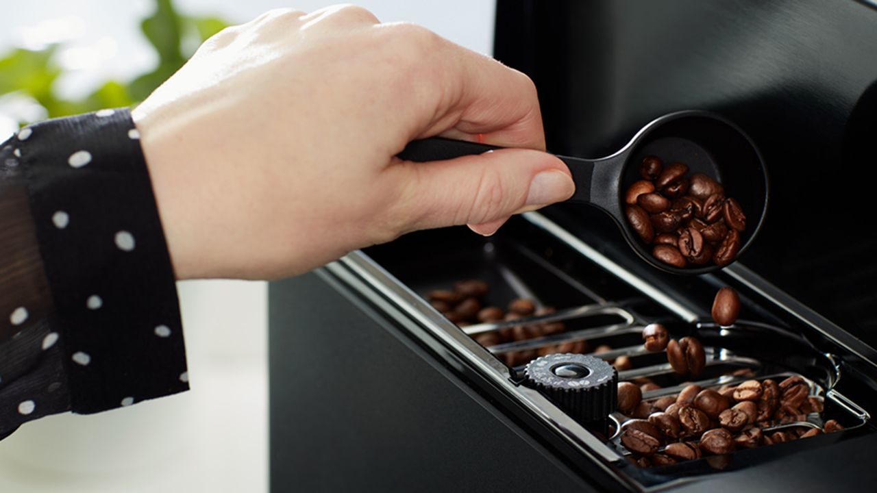 A female wearing a black blouse using a scoop with whole coffee beans demonstrating how to use a coffee maker