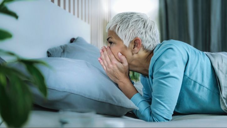 A woman with short grey hair wearing a blue pj top struggling to sleep during menopause. She lays on her front with hands over her mouth and nose in bed with grey bedding