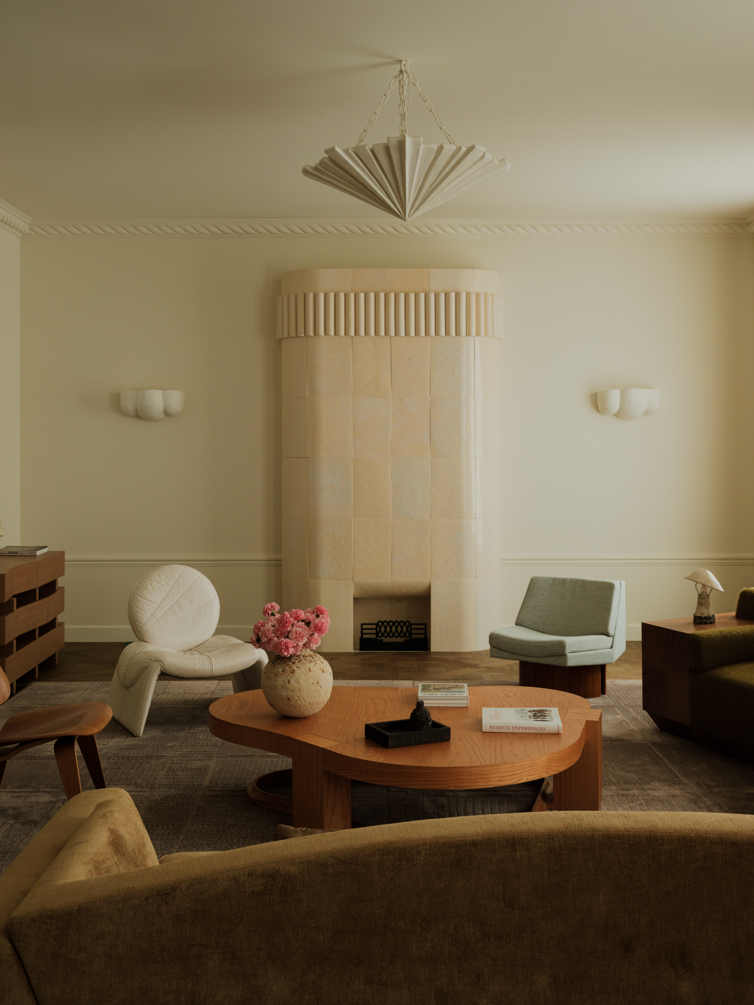 Image of a soft minimalist living room with white walls and curved shapes. There is a pastel stone, rounded fireplace, a curved wooden coffee table, and a mix of accent chairs around the room.