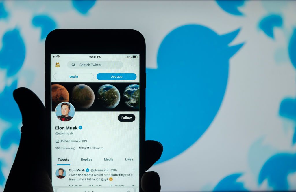 Elon Musk&amp;#039;s twitter account is seen displayed on a mobile phone screen with a twitter logo in the background.