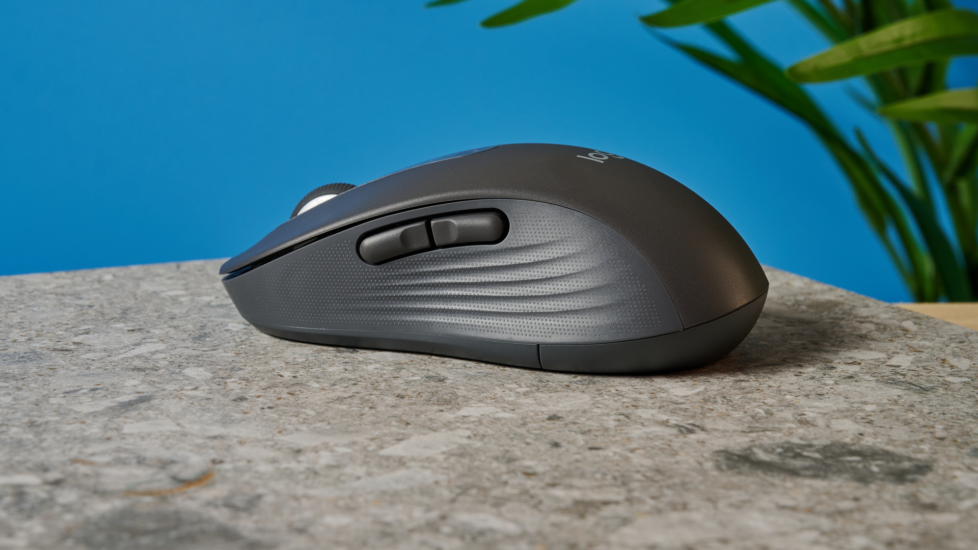 a black mouse with a scroll wheel, two side buttons, and grooved edges, with a logi logo, sits atop a pale table with a blue background