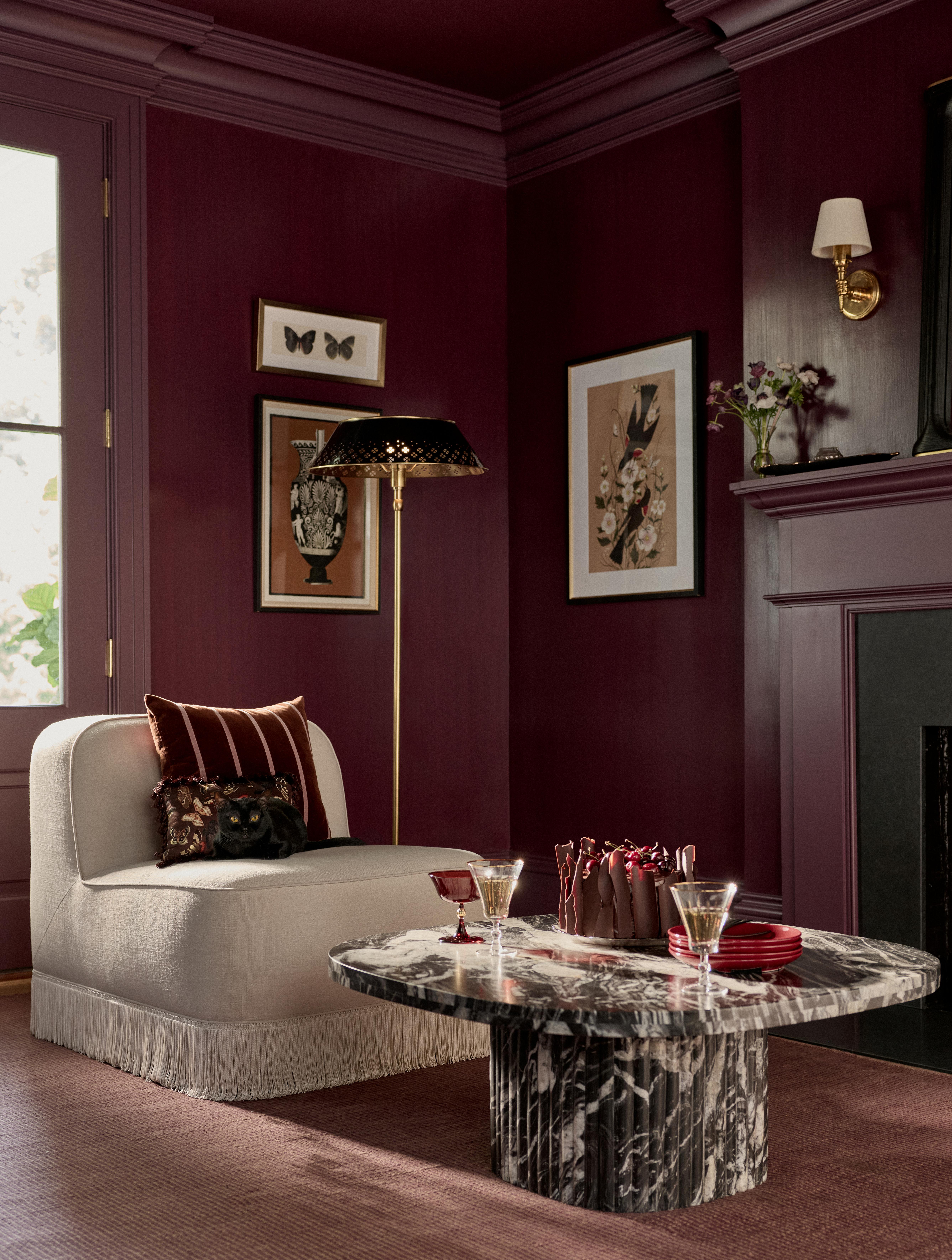 Anthropologie x Catherine Martin Halloween collection, including tableware and framed art prints, photographed in a burgundy living room with a marble coffee table and fringe slipchair