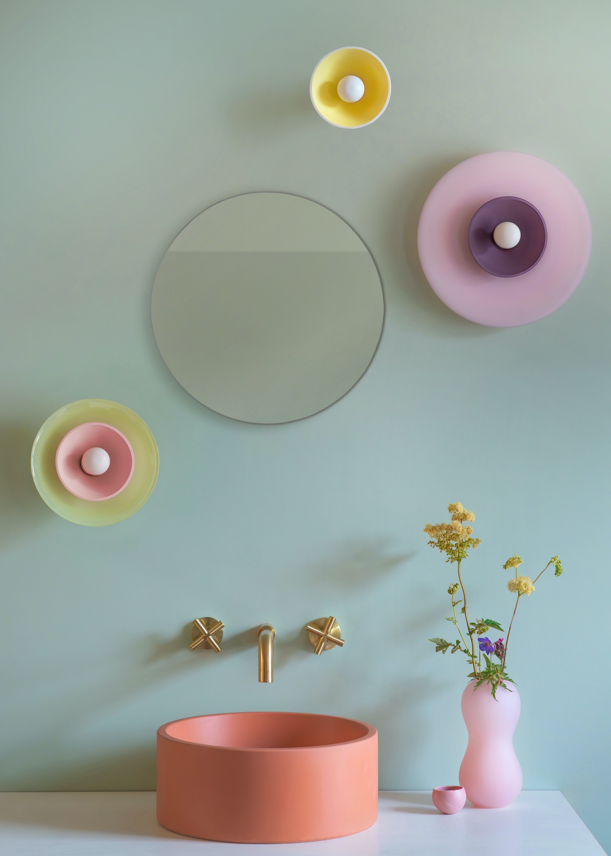 colorful wall lights with orange basin and brass taps and a round mirror and pink vase of yellow flowers