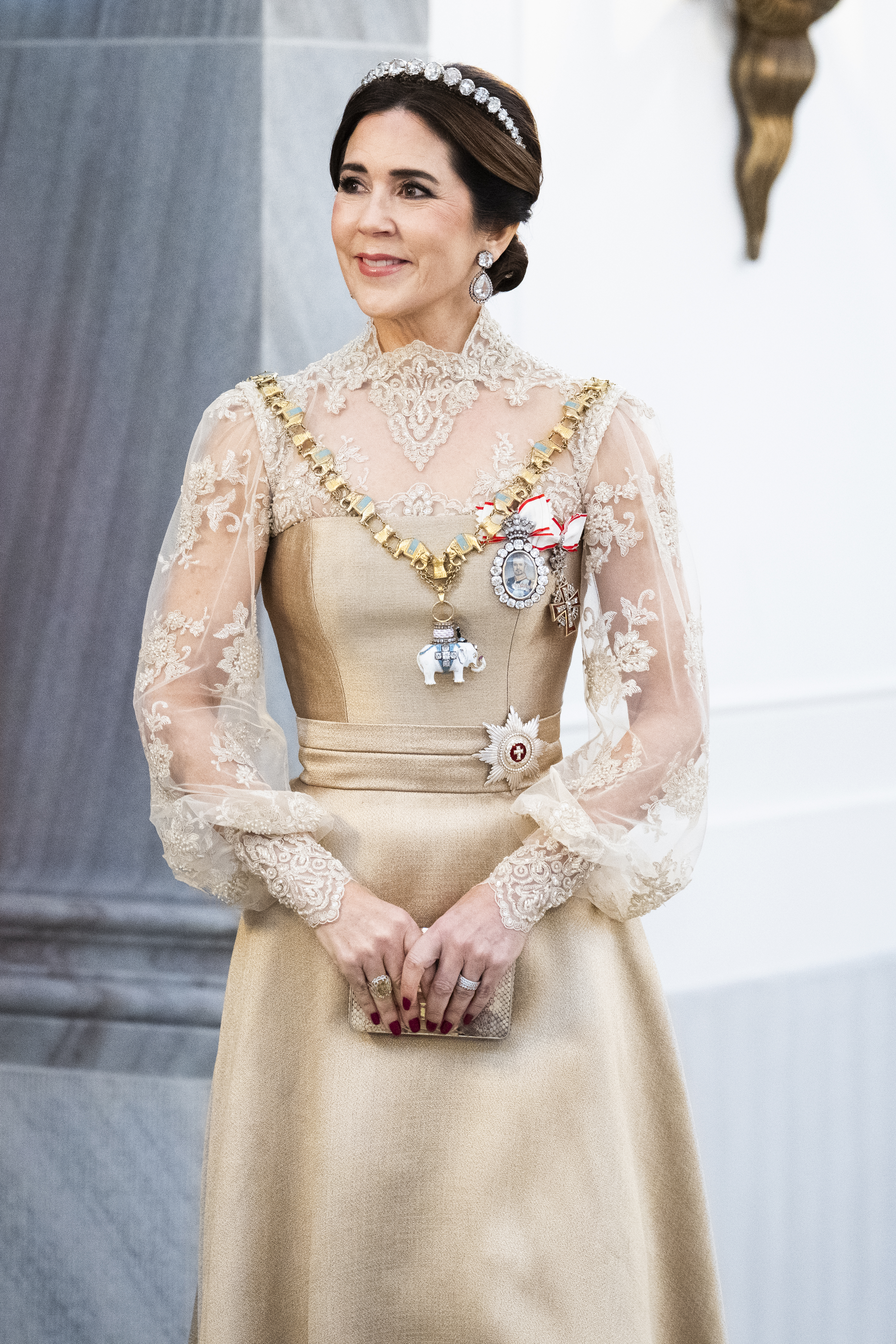 Queen Mary wearing a gold gown and diamond tiara standing with her arms in front