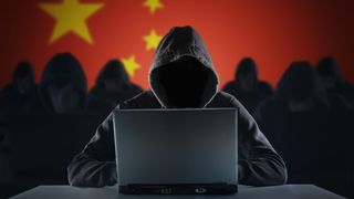 A group of 7 hackers, 6 slightly blurred in the background and one in the foreground, all wearing black with hoods pulled up over their heads. You cannot see their faces. The hacker in the foreground sits with an open laptop in front of them. The background, behind the hackers, is a Chinese flag