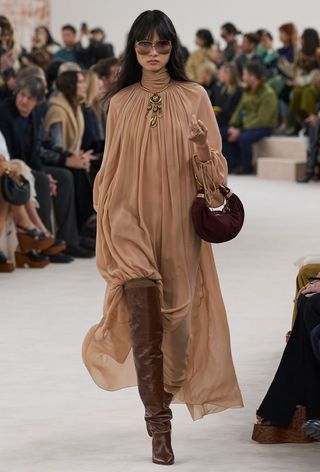 a photo showing some of the best thigh-high boots with a model walking the Chloé runway show wearing a ruffled tan turtleneck dress with brown thigh-high boots, a red suede bag, and a gold necklace