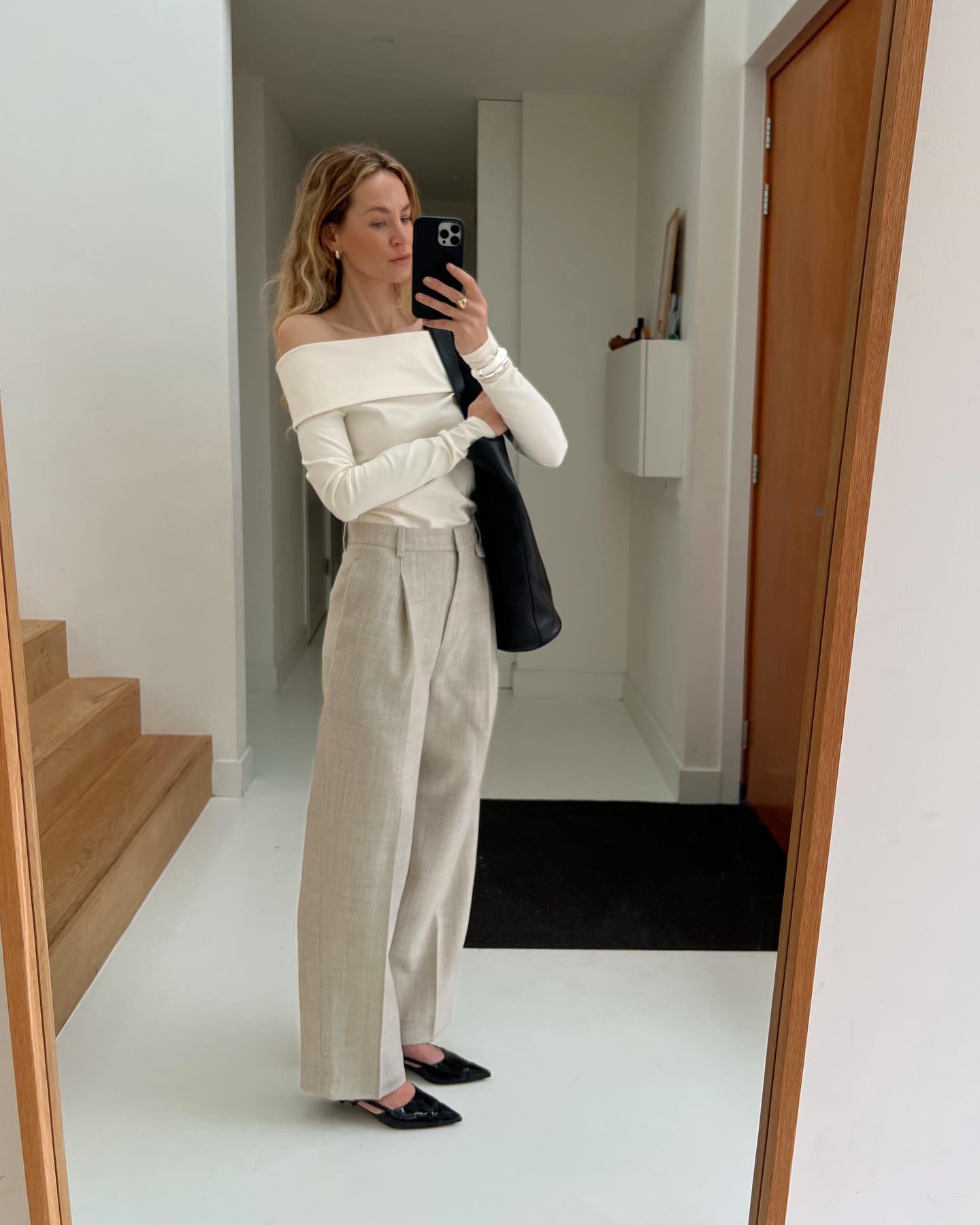 fashion influencer Anouk Yve poses for a mirror selfie wearing a cream off-shoulder top, oversize black tote bag, tan linen pants, and black slingback heels
