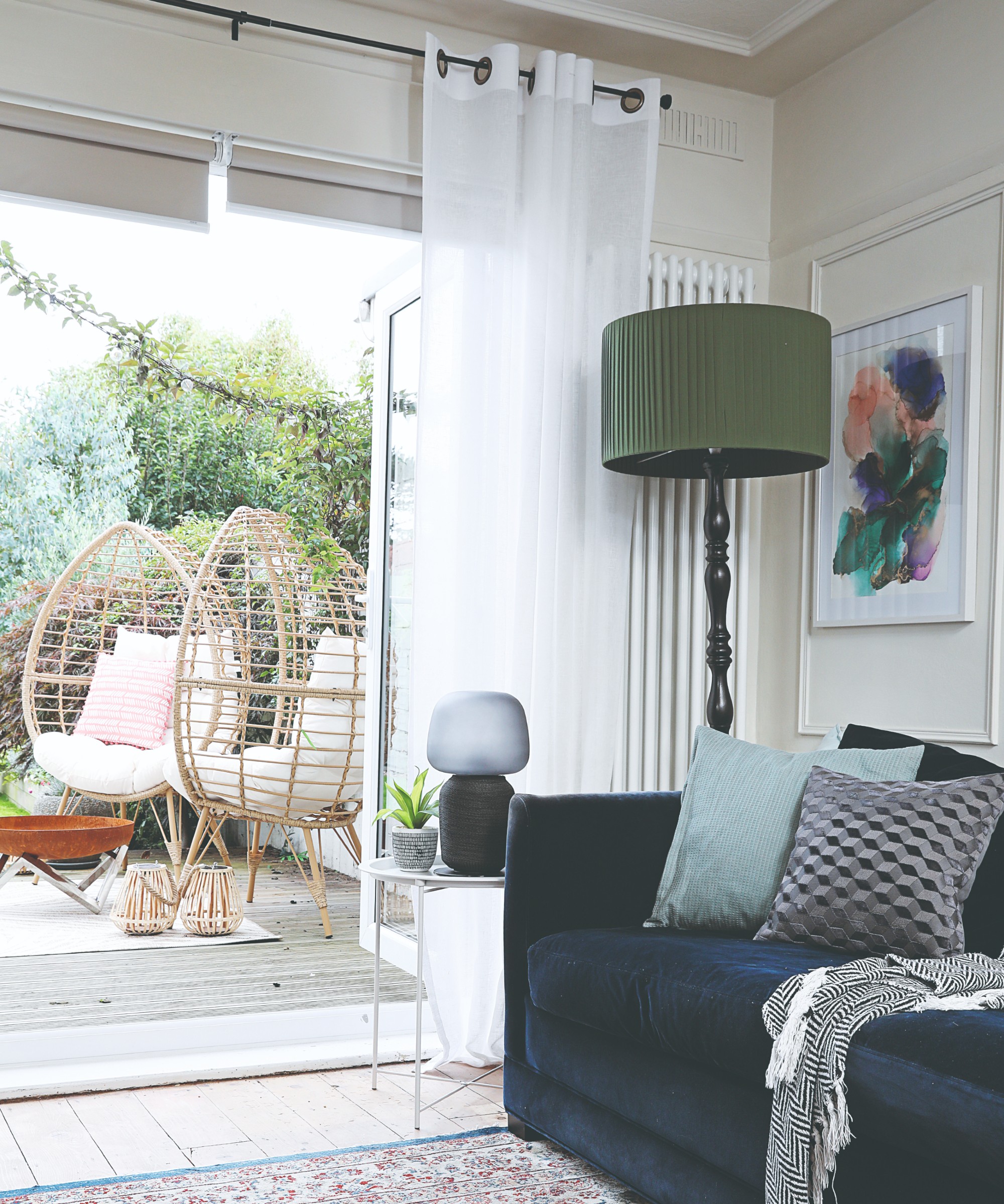 A living room with a blue velvet sofa and glass doors open to the garden dressed with white voile curtains and roller blinds