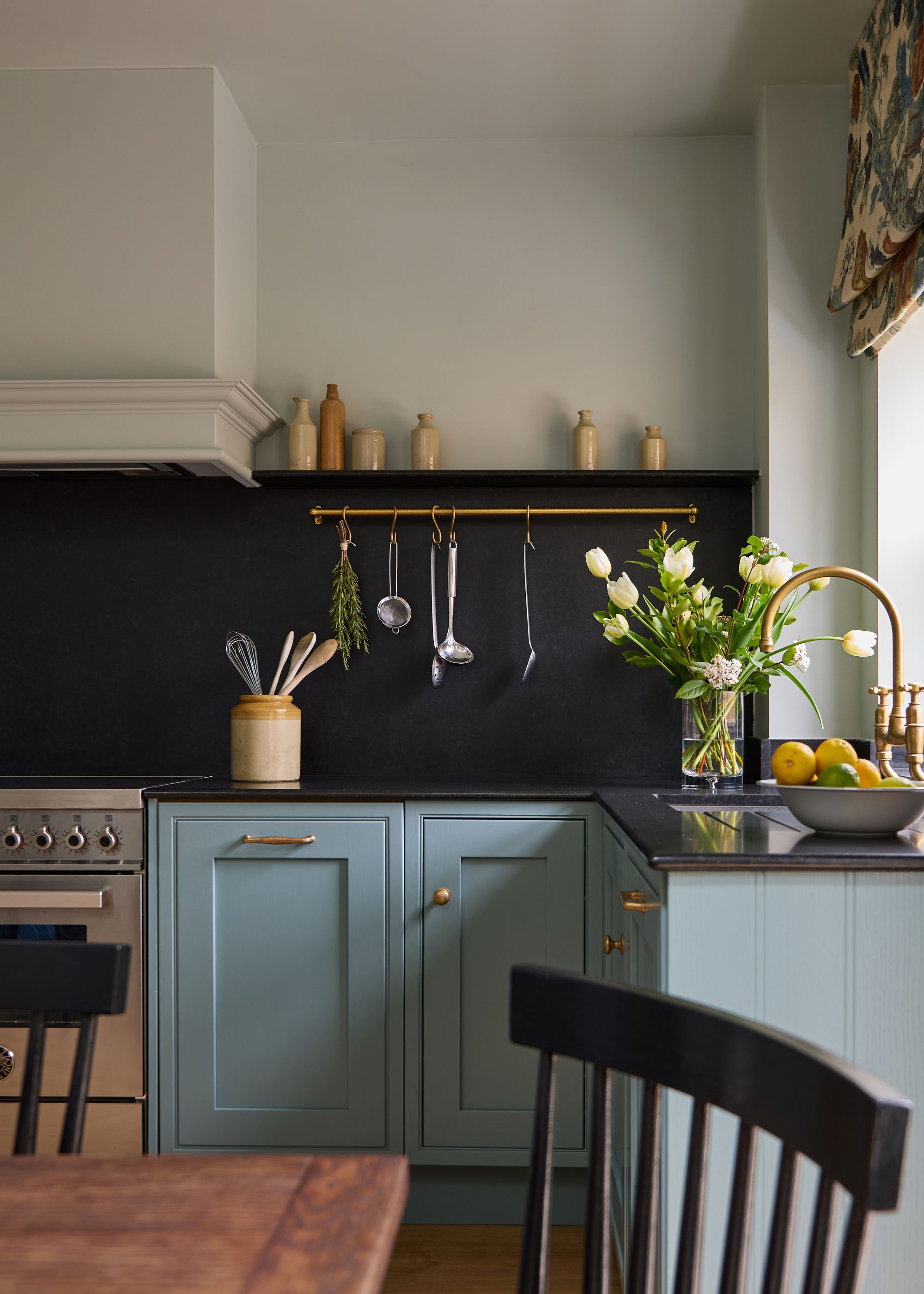 A kitchen with blue-green lower cabinets, a black countertop and backsplash, warm white walls, brass fixtures, and a brass rail for utensils.