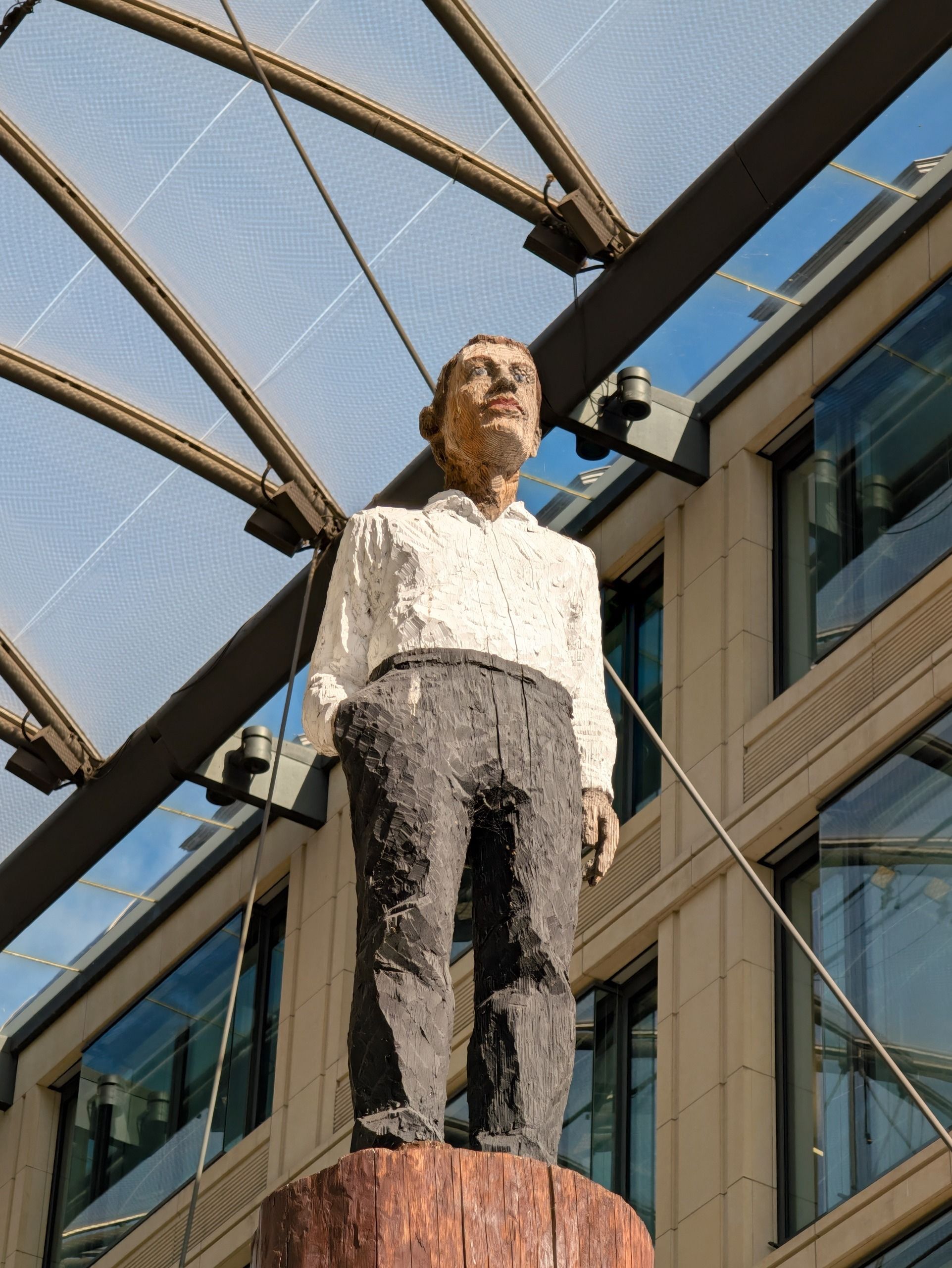 Tall carved wooden sculpture of a man in a white shirt and dark trousers beneath a glass canopy.