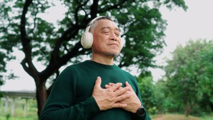 An older man outside, wearing headphones and with his hands place on his chest, breathes in as if relaxing.