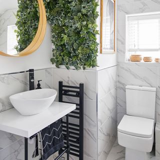 downstairs toilet with white toilet and sink, circular mirror and faux plant wall
