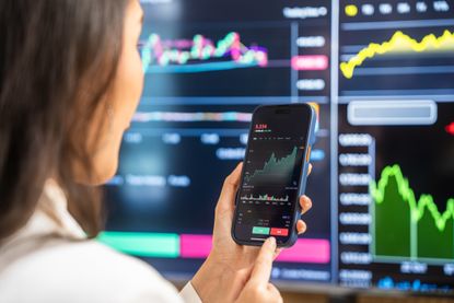 A businesswoman examines financial charts and graphs on her smartphone, utilizing modern technology for investment analysis amidst digital screens displaying stock data.