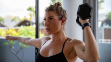 A woman performs a home dumbbell workout, holding a dumbbell in one hand at shoulder height while the other arm remains extended to the side. 