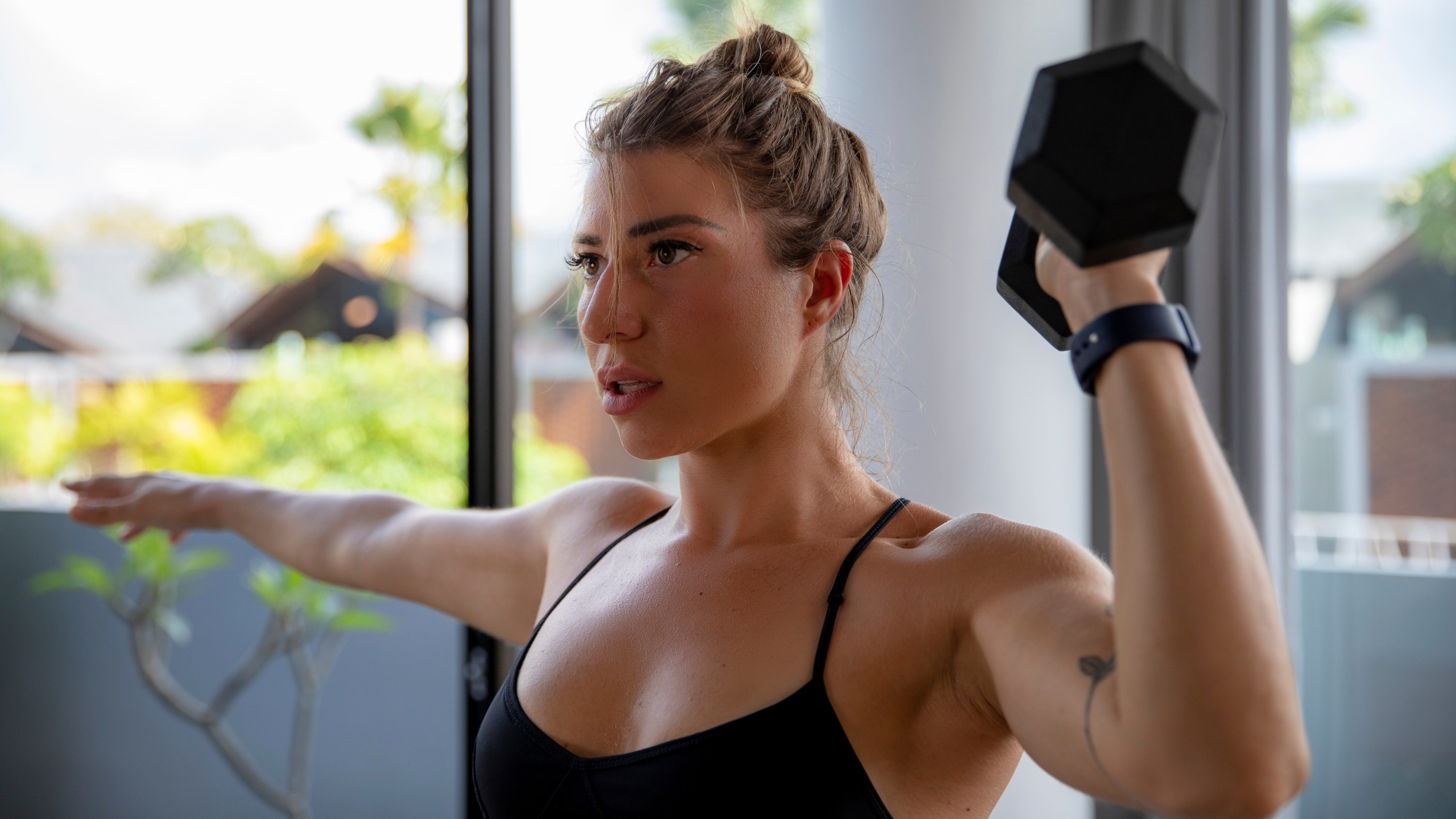 A woman performs a home dumbbell workout, holding a dumbbell in one hand at shoulder height while the other arm remains extended to the side. 