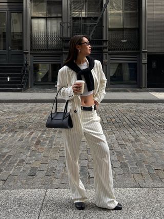 a photo of a woman wearing a white-and-black pinstripe suit with a white tank top, black cardigan, black shoulder bag, and ballet flats