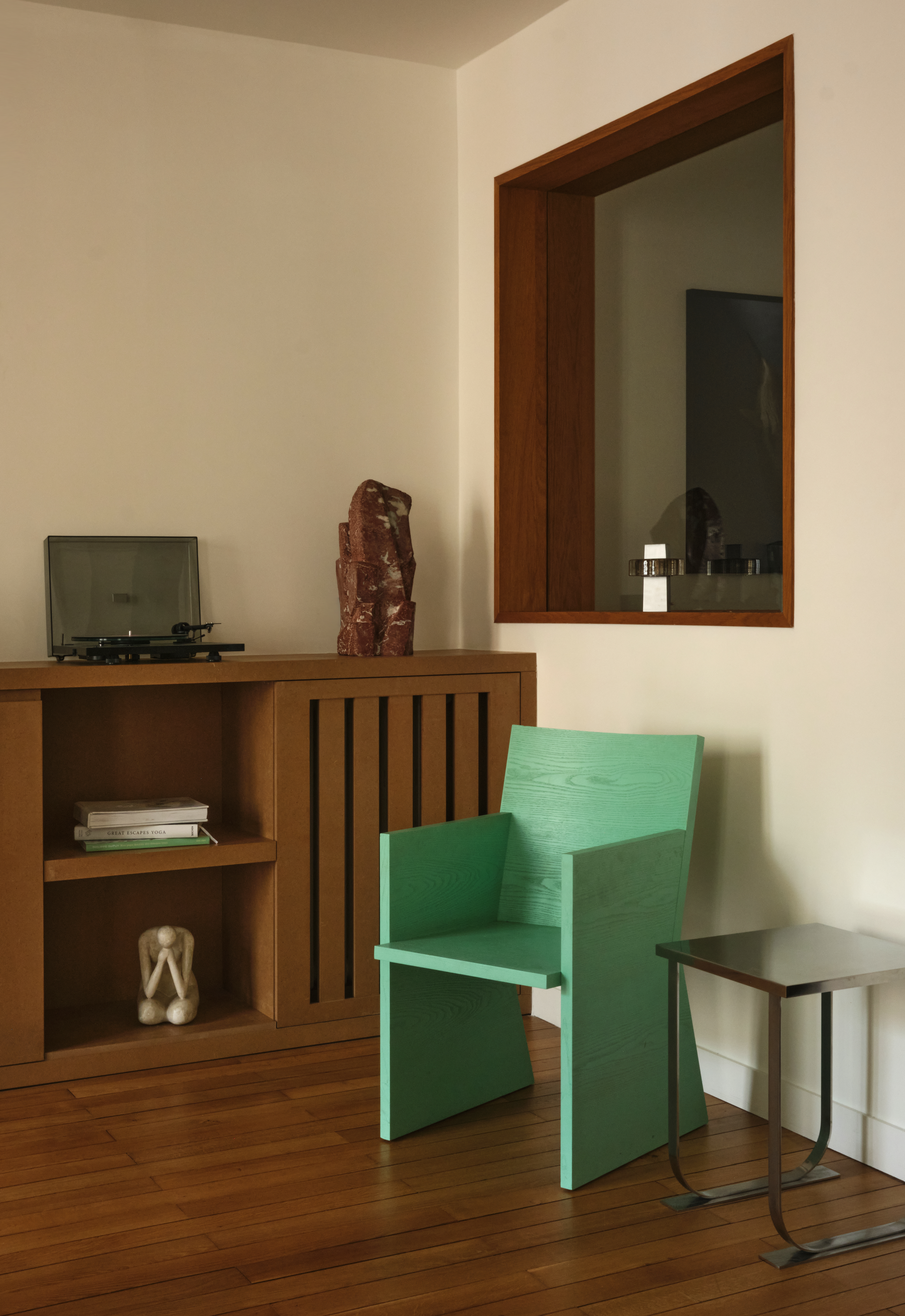 A marine green sculptural chair, placed in a naturally lit living room with wooden floors and furniture.