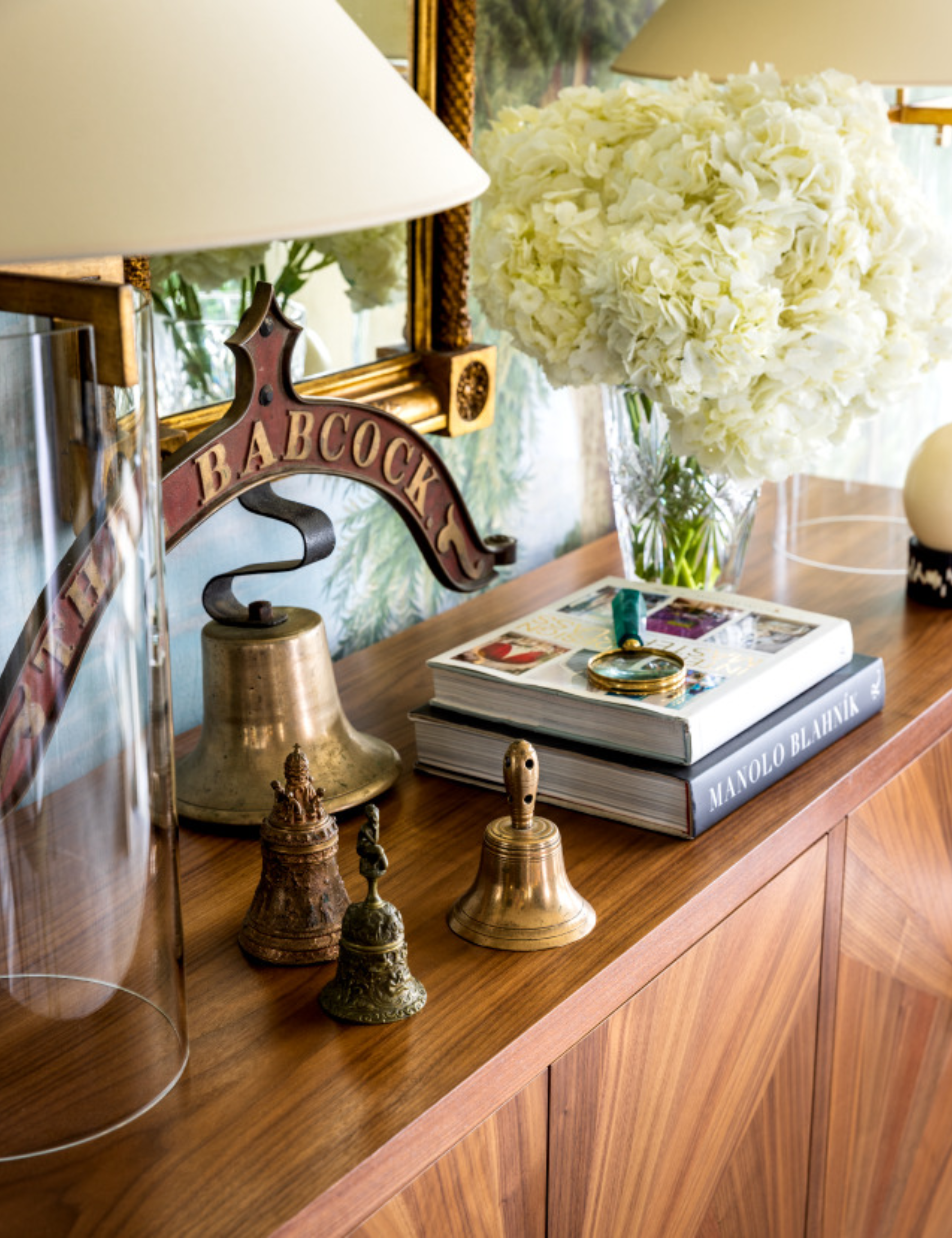 Wooden sideboard topped with vintage brass bells, a bookstack, table lamps, and a vase full of hydrangeas