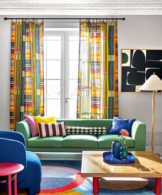 Colorful, contemporary living room with abstract patterned curtains, gray painted walls, green sofa, blue armchair, colorful rug, wooden square coffee table with red feet