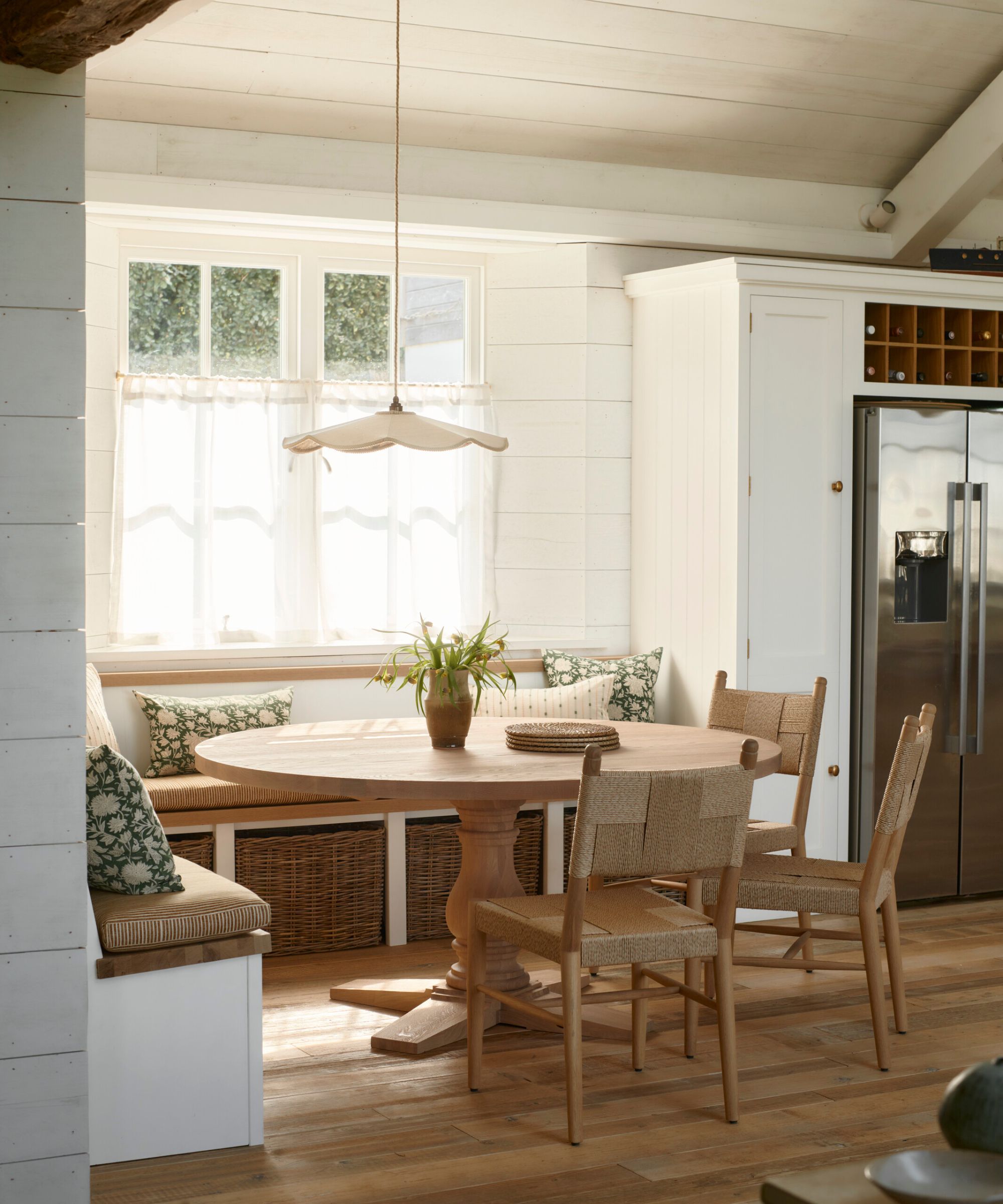 A white coastal kitchen with a dining nook in the corner made up of a corner banquette and a round wooden table