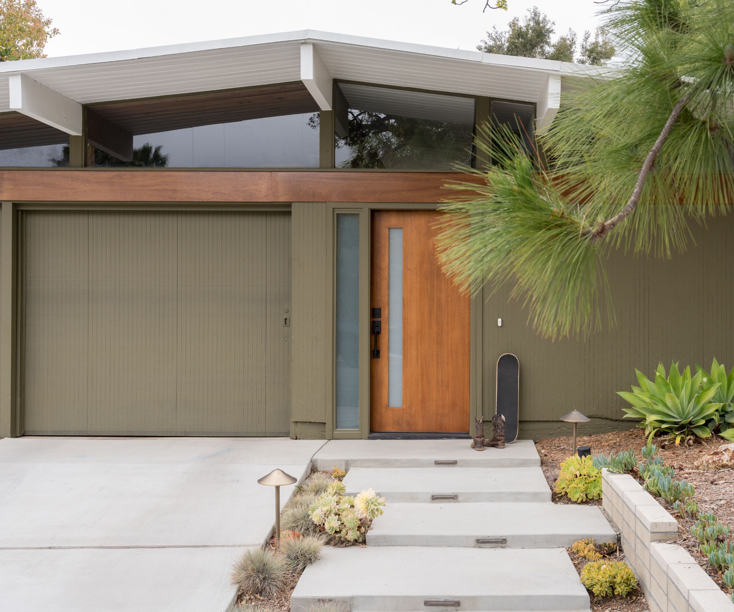 A modern home with olive green exterior, a sloped white roof, a glass upper wall, and a wooden front door with a modern front yard.