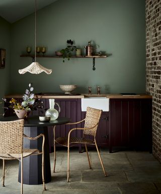 Rustic kitchen-diner with green painted wall, exposed brick wall, rounded dining table with two wicker dining chairs, low hanging scalloped style pendant light