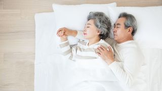 ¿Cuánto sueño necesitas? Una pareja mayor durmiendo de lado acurrucados en una cama blanca.