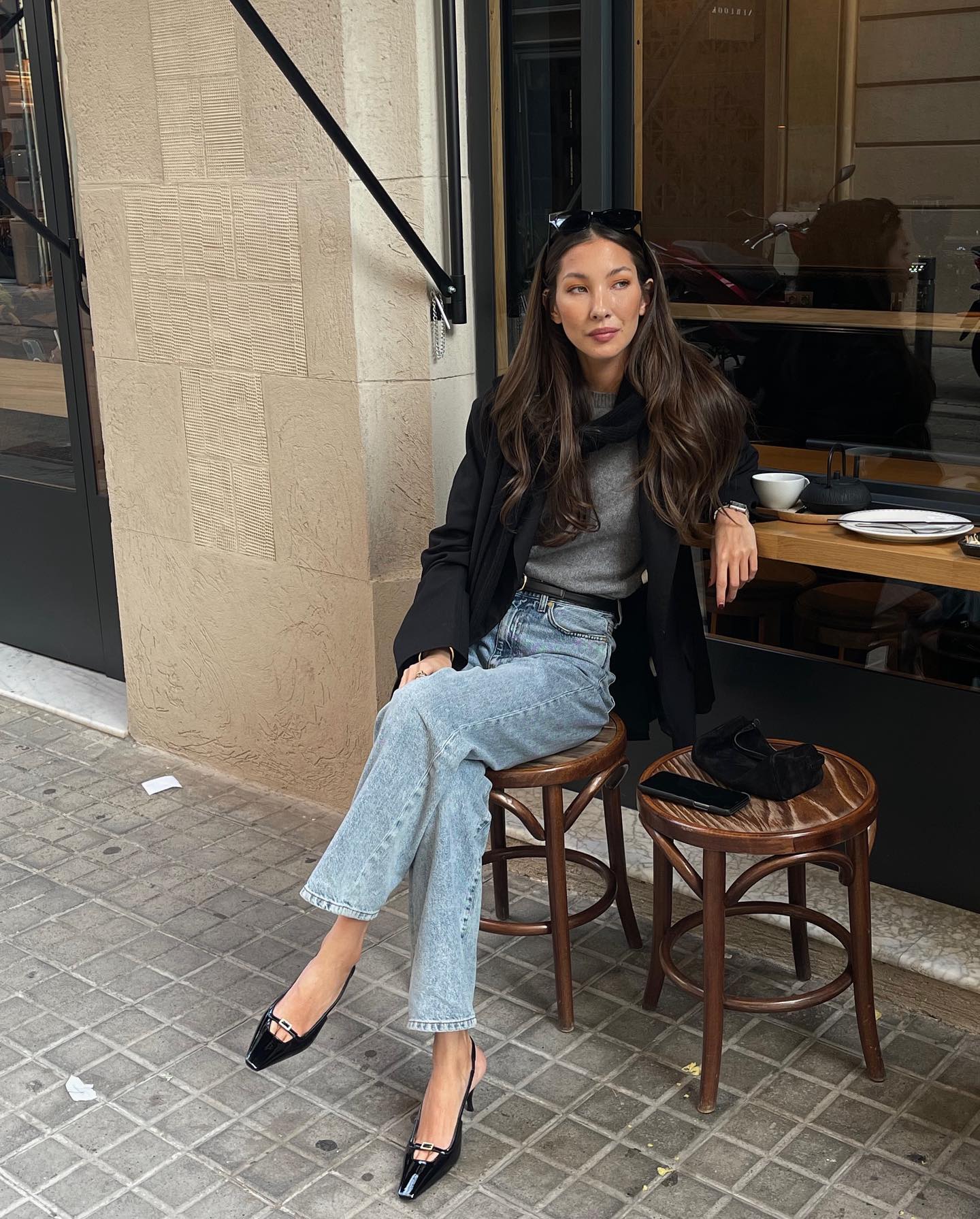 fashion influencer Felicia Akerstrom sits at a cafe in a black blazer, gray sweater, jeans, and black slingback heels