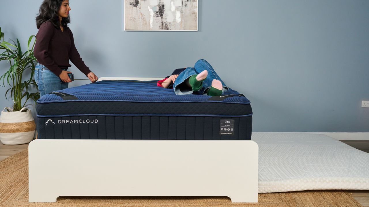 The image shows dark haired mattress tester standing next to a dark blue mattress with a measuring tape. On the opposite side of the bed, a second mattress tester slowly rolls off the side. This roll off test determines how sturdy the bed's edge is.
