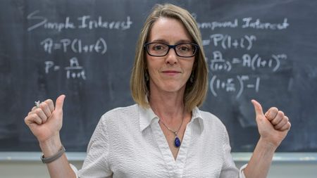 A woman stands in front of a chalkboard with a math formula on it.