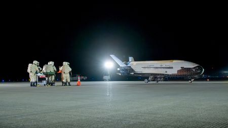 technicians in bulky protective suits approach a robotic winged space plane at night