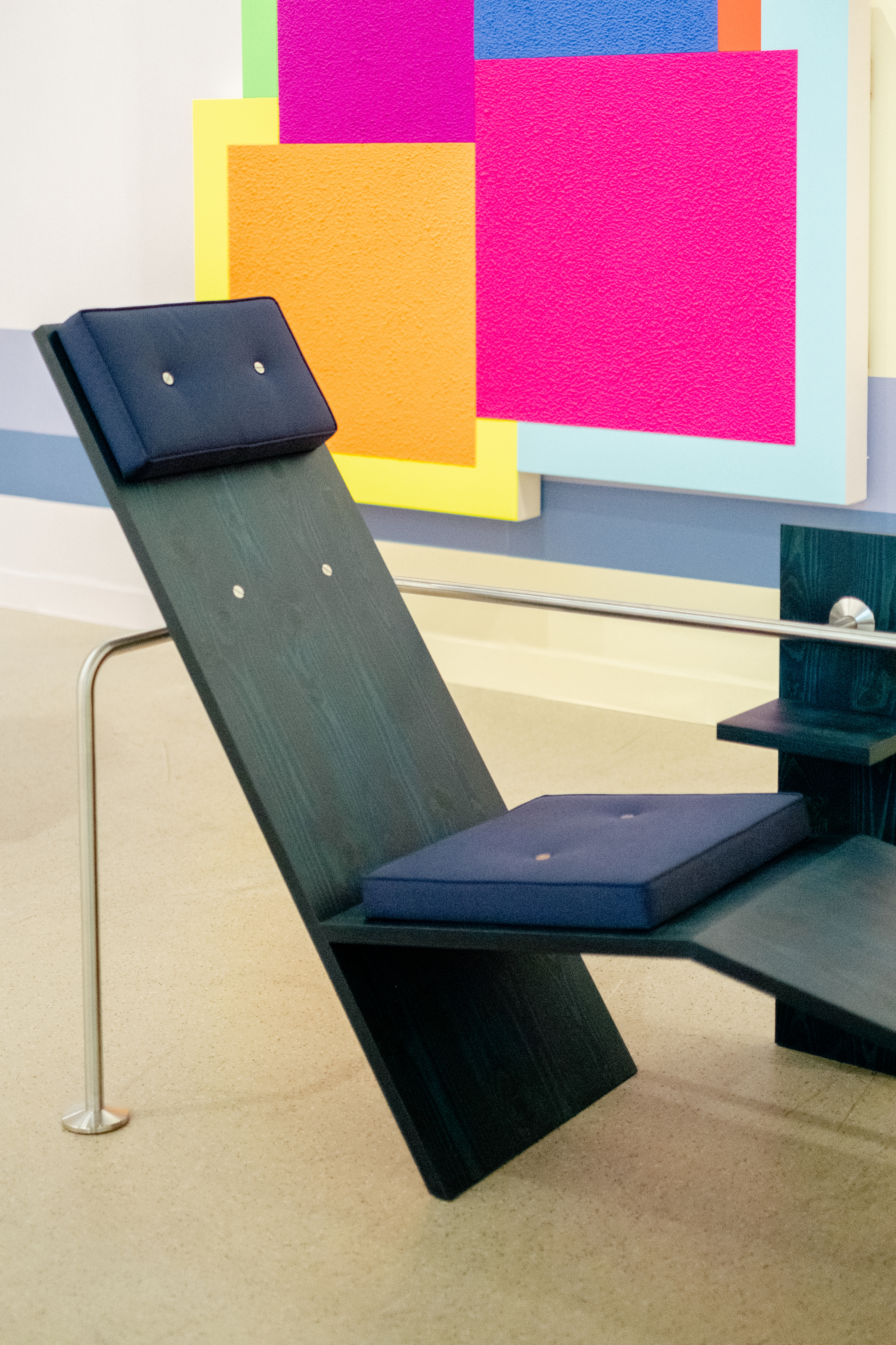 A modernist-inspired wood, chrome, and textile chair features a minimalist, playful design while sitting against a colorful, abstract artwork.